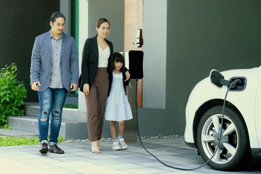 Progressive young parents and daughter with electric vehicle and home charging station. Green and clean energy from electric vehicles for healthy environment. Eco power from renewable source at home.