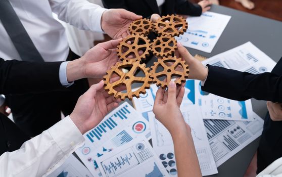 Closeup top view businesspeople hand holding gear and join together over meeting table with financial report papers. Cohesive group of office workers holding cog wheel as synergy harmony concept.