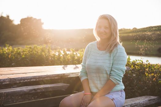 blonde girl in shorts sitting at sunset at the wooden table near the lake