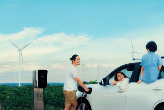 Concept of progressive happy family enjoying their time at wind farm with electric vehicle. Electric vehicle driven by clean renewable energy from wind turbine generator for charging station.