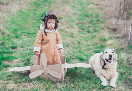 Charming baby in aviator's clothes and his co-pilot are standing by a cardboard plane