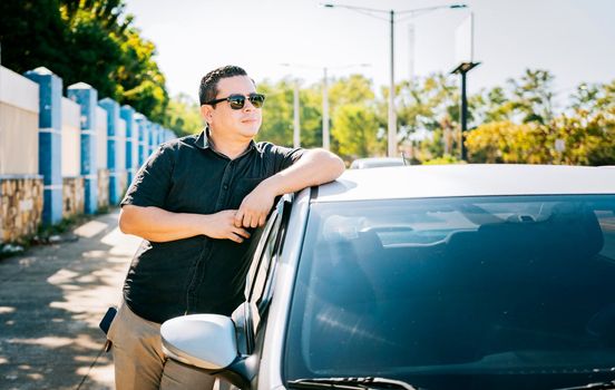 Smiling driver leaning on the car door. Smiling owner with new car. Young latin man leaning on car door in the street, Happy driver leaning on the car door in the street