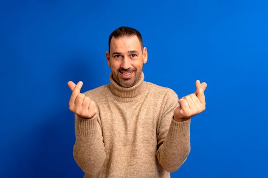 Bearded latino man wearing a turtleneck sweater making the money gesture with his fingers isolated on blue studio background