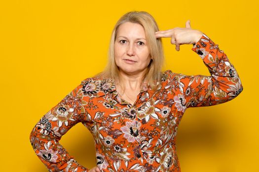 Caucasian blonde woman in a patterned dress making the gesture of a gun pointing at her head isolated over yellow background