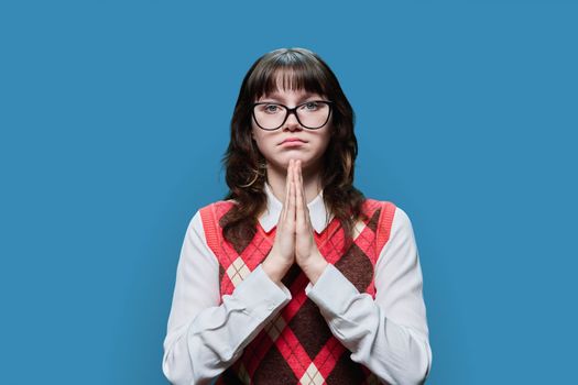 Young sad female in glasses pleading holding her palms together, on blue studio background. Girl student looking at camera, in request, in plea, asking for forgiveness for help sorry pray please