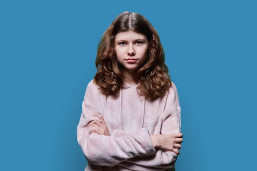Portrait of calm serious child schoolgirl on blue studio background. Preteen girl looking at camera with crossed arms, wearing school clothes. School, kids, study education, childhood concept