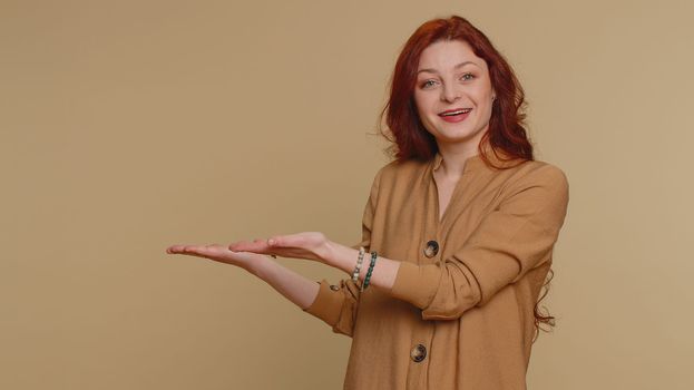 Lovely millennial redhead woman showing thumbs up and pointing empty place, advertising area for commercial text, copy space for goods promotion. Young girl. Studio shot indoors on beige background