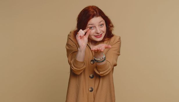 Need some more, please give me. Redhead woman showing a little bit gesture with sceptic smile, showing minimum sign, measuring small size. Young ginger girl isolated alone on beige studio background