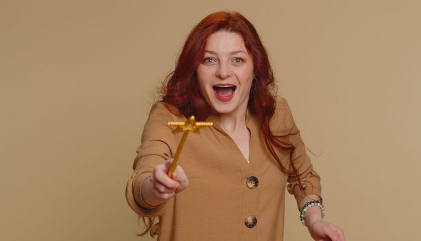 Magician witch woman gesturing with magic wand fairy stick, making wish come true, casting magician spell, advertising holidays sale discount. Young redhead girl isolated on beige studio background