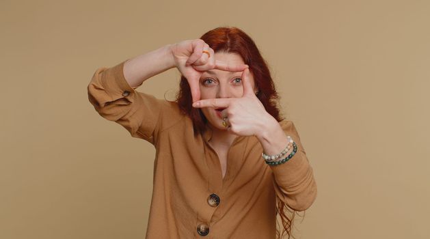 Redhead woman photographer gesturing picture frame with hands, looks through fingers and focusing on interesting moment, imitating zoom and cropping nice image. Young girl on beige studio background