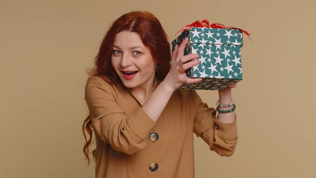 Happy redhead woman opening gift box, unwrapping birthday surprise and expressing great happiness amazement, satisfied with best present bonus. Young ginger girl isolated on beige studio background