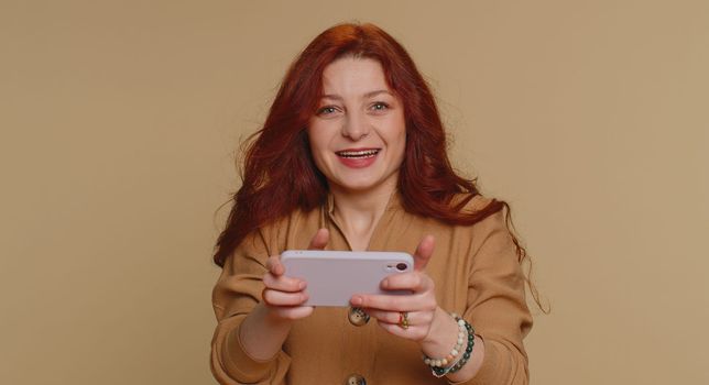 Worried funny addicted woman enthusiastically playing racing video games on mobile phone. Young redhead girl using smartphone gadget app with drive simulator isolated on beige studio background