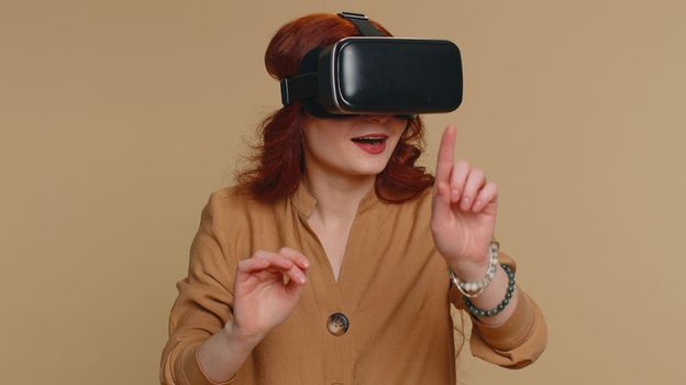Redhead woman using headset helmet to play simulation game app online. Watching virtual reality 3D 360 video content. One girl in VR goggles isolated on studio beige background. Futuristic technology