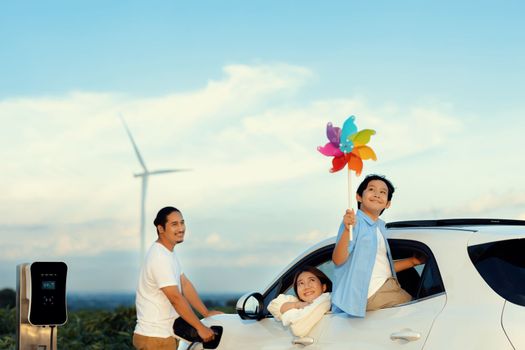 Concept of progressive happy family enjoying their time at wind farm with electric vehicle. Electric vehicle driven by clean renewable energy from wind turbine generator for charging station.