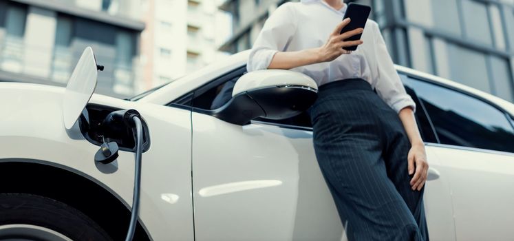 Closeup progressive businesswoman with smartphone, leaning electric car and charging station before driving around city center. Eco friendly rechargeable EV car powered by sustainable and clean energy