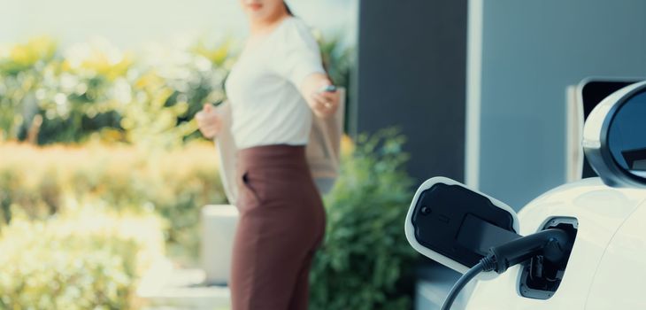 Focus EV charger plugged into EV car at home charging station with blurred background of progressive woman walking in background. Elective vehicle powered by clean energy for eco-friendly concept.