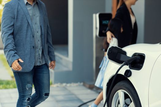 Closeup focus hand insert EV charger plug to electric vehicle at home charging station with blurred family in background. Progressive concept of green and clean energy to reduce CO2 emission by EV car