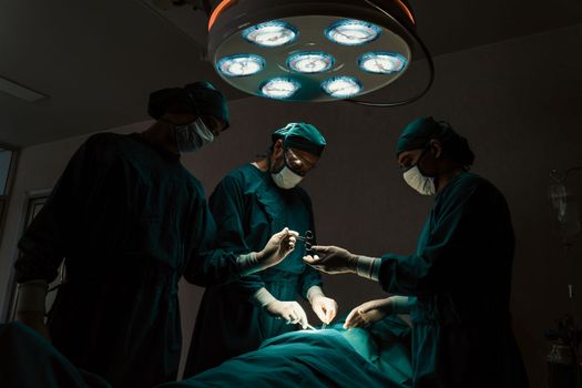 Surgical team performing surgery to patient in sterile operating room. In a surgery room lit by a lamp, a professional and confident surgical team provides medical care to an unconscious patient.