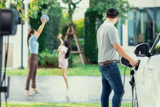 Focus image of progressive man charging electric car from home charging station with blur mother and daughter playing together in the background.