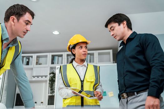 A team of competent engineers wearing safety equipment is working on blueprints with a tablet while also discussing with the investor in the office.