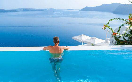 man relaxing in infinity swimming pool during vacation at Santorini, swimming pool looking out over the Caldera ocean of Santorini, Oia Greece, Greek Island Aegean Cyclades luxury vacation.