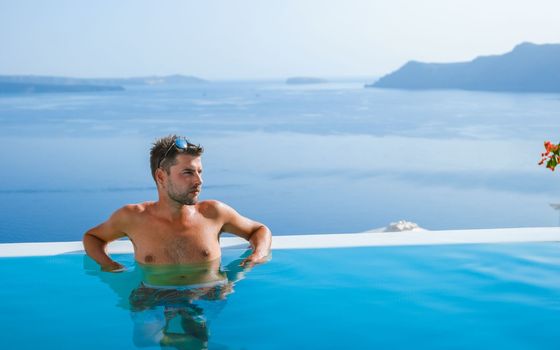 man relaxing in infinity swimming pool during vacation at Santorini, swimming pool looking out over the Caldera ocean of Santorini, Oia Greece, Greek Island Aegean Cyclades luxury vacation.