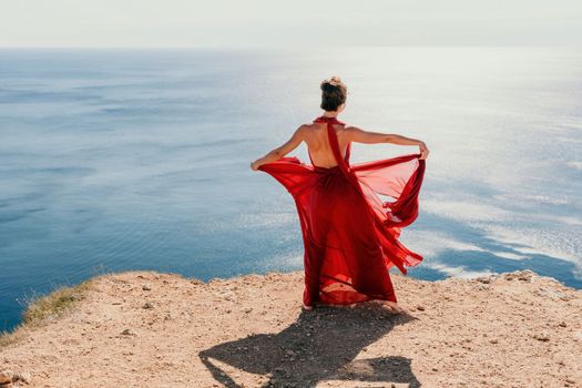 Side view a Young beautiful sensual woman in a red long dress posing on a rock high above the sea during sunrise. Girl on the nature on blue sky background. Fashion photo.