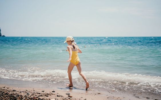 Cute little girl running along the seashore against a clear blue sea and rejoices in the rays of the summer sun. Beautiful girl in yellow swimsuit running and having fun on tropical beach