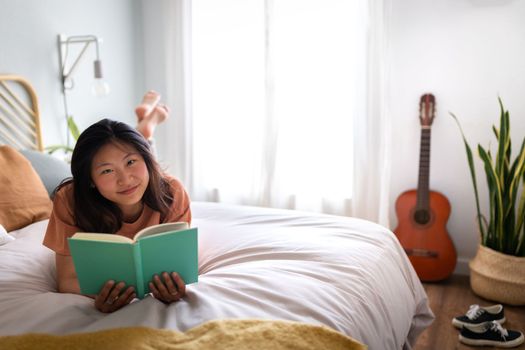 Smiling Asian young woman reading book lying on bed, relaxing in cozy bedroom looking at camera. Copy space. Comfort, leisure and home concepts.