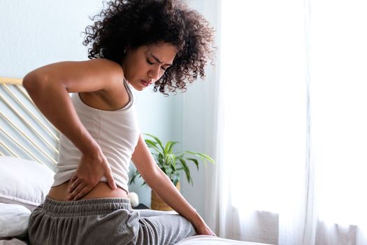 Rear view of young african american woman touching lumbar feeling back pain. Female sitting on bed with back ache. Copy space. Body concept.