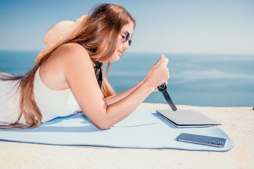 Successful business woman in yellow hat working on laptop by the sea. Pretty lady typing on computer at summer day outdoors. Freelance, travel and holidays concept.