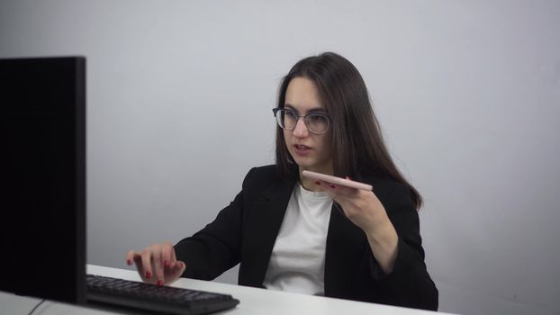 A young businesswoman is recording a voice message while sitting in the office. A woman in glasses and a suit at the workplace with a phone in her hands. 4k