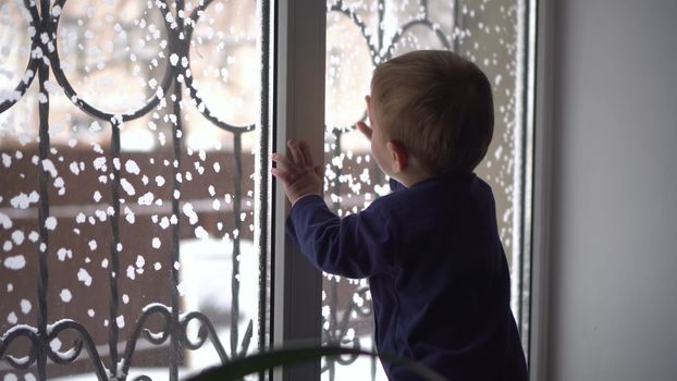 A small child stands on the windowsill and looks out the window. A child knocks on glass in winter. 4k