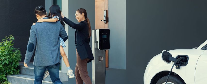 Progressive young parents and daughter with electric vehicle and home charging station. Green and clean energy from electric vehicles for healthy environment. Eco power from renewable source at home.