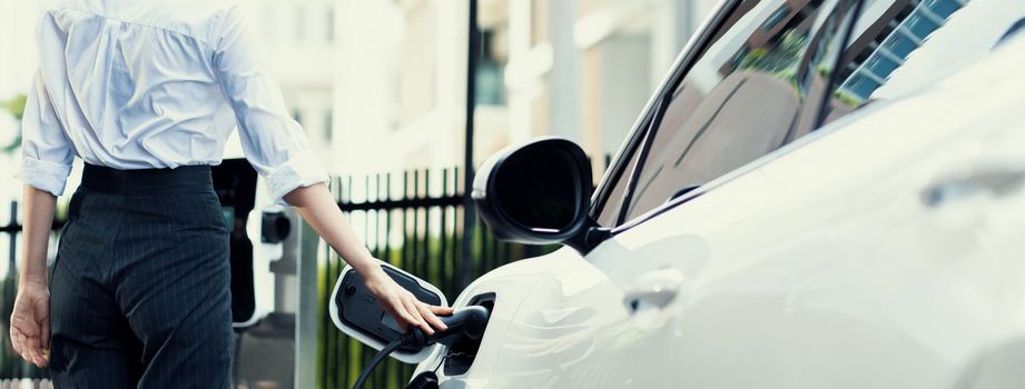 Closeup progressive suit-clad businesswoman with her electric vehicle recharge her car on public charging station in modern city with power cable plug and renewable energy-powered electric vehicle.