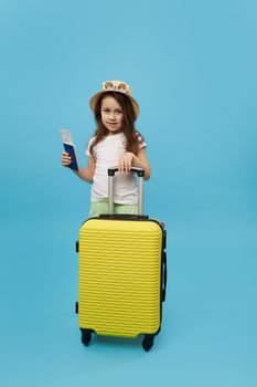 Full length portrait of a little girl in summer wear, holding a flight ticket, standing by a bright yellow suitcase, smiles, looking at camera, isolated blue background. Happy summer holidays concept