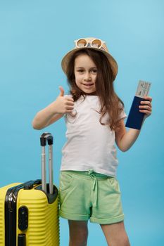 Adorable lovely 5 years little traveler girl in summer wear, showing thumb up looking at camera, posing with a flight ticket , boarding pass and yellow suitcase, blue color background. Trip and travel
