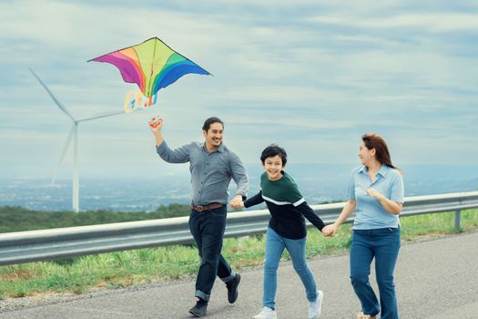 Progressive happy carefree family vacation concept. Young parents mother father and son run along and flying kite together on road with natural scenic on mountain and wind turbine background.