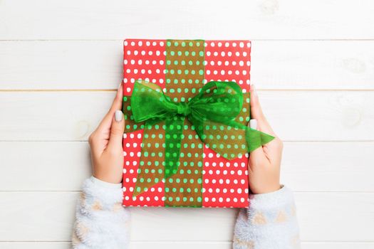 Woman hands give wrapped christmas or other holiday handmade present in colored paper . Present box, decoration of gift on white wooden table, top view with copy space.