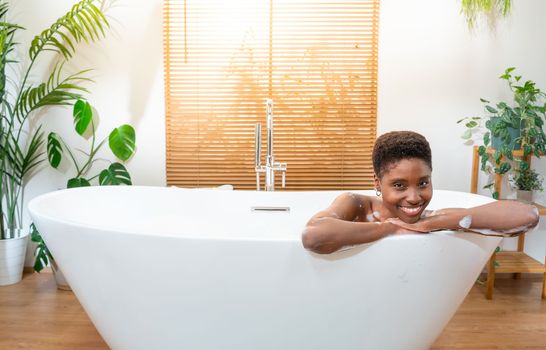 Luxury bath portrait of an attractive African woman relaxing in bathtub at cozy home bathroom looking at camera smiling happy. Real people wellness concept. High quality photo