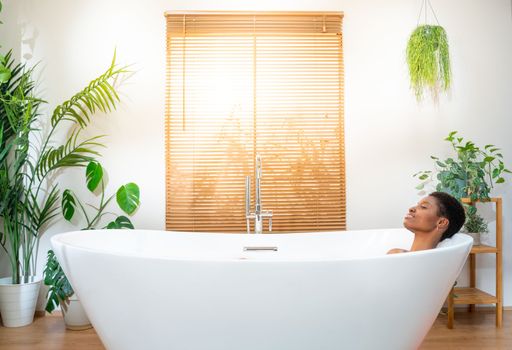 Attractive woman relaxing in foam bath in beautiful bathroom with plants at home during a sunny morning routine. Rest, calm, pleasure, happiness and wellbeing concept. High quality photo