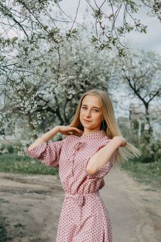 Blonde girl on a spring walk in the garden with cherry blossoms. Female portrait, close-up. A girl in a pink polka dot dress