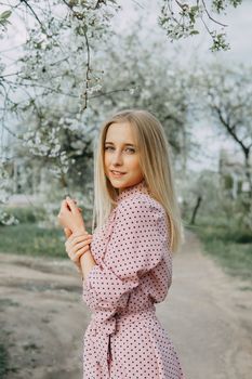 Blonde girl on a spring walk in the garden with cherry blossoms. Female portrait, close-up. A girl in a pink polka dot dress