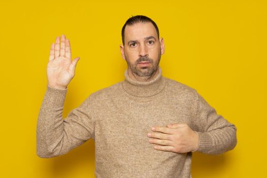 Bearded handsome man wearing turtleneck sweater standing over isolated yellow background. Swearing with hand on chest and open palm, swearing a pledge of allegiance