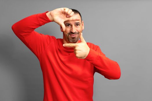 Handsome bearded happy male blinks his eyes and looks through a frame formed by his hands. An attractive man makes a frame with his fingers, isolated on a gray background with copy space for text.