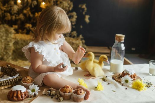 A little girl is sitting on the Easter table and playing with cute fluffy ducklings. The concept of celebrating happy Easter