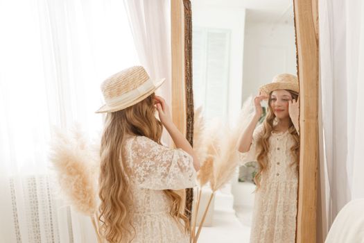 A beautiful teenage girl with long hair measures a straw hat in front of a mirror. Self-admiration of a blonde. selective focus
