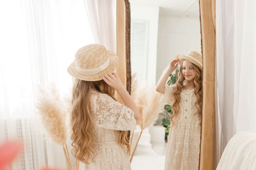 A beautiful teenage girl with long hair measures a straw hat in front of a mirror. Self-admiration of a blonde. selective focus