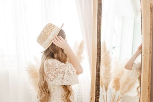 A beautiful teenage girl with long hair measures a straw hat in front of a mirror. Self-admiration of a blonde. selective focus