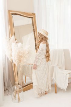 A beautiful teenage girl with long hair measures a straw hat in front of a mirror. Self-admiration of a blonde. selective focus